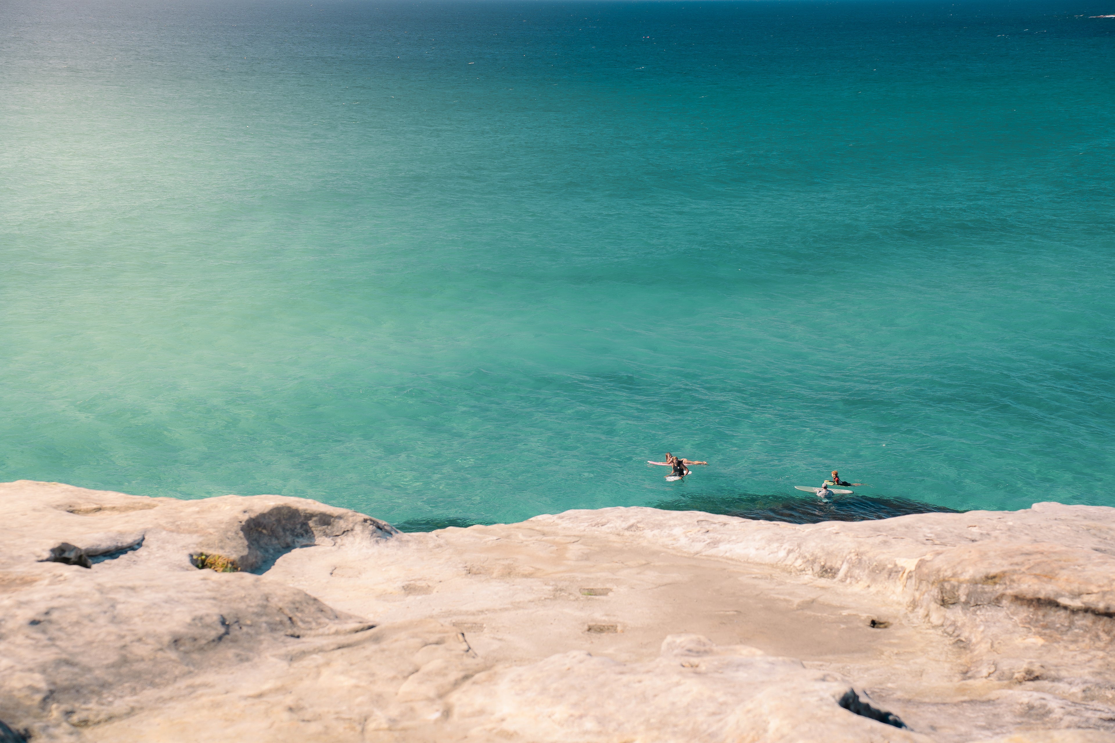 Four people on surfboards on a turquoise sea from a high vantage point.